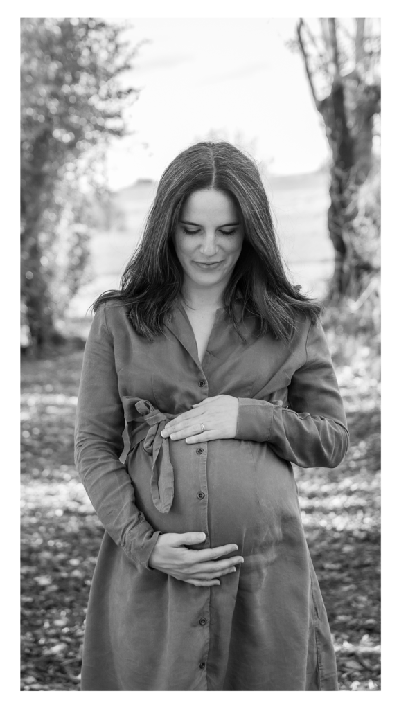 Portrait en noir et blanc de la future maman — regard vers le bas, mains sur le ventre, arbres en fond. Douceur et intériorité.